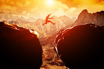 Man jumping over precipice between two mountains at sunset